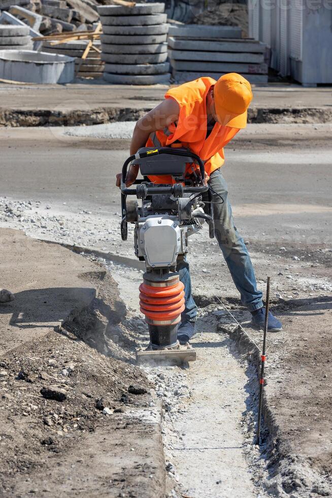 A builder in an orange cape is ramming a trench with a vibratory rammer at a construction site. photo