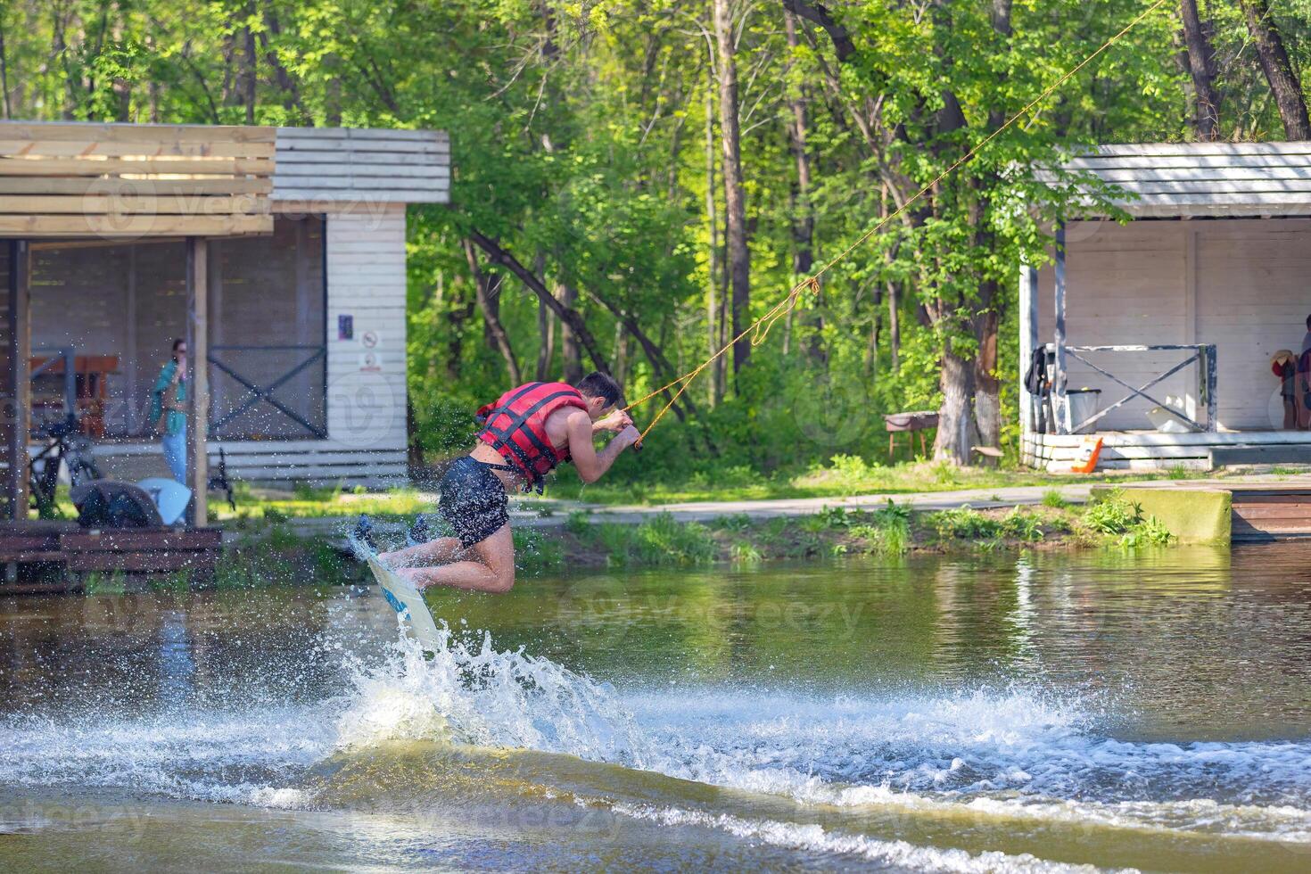 Wakeboarder leaps from the water, creating splashes under the bright sun in vibrant summer day of wakeboarding fun at the lakeside park. photo
