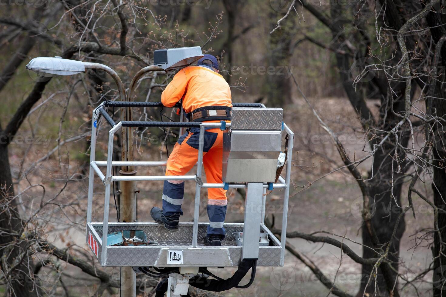 Worker in bright orange safety suit on elevated platform fixing streetlight amidst bare trees, maintenance task, outdoor scene concept photo
