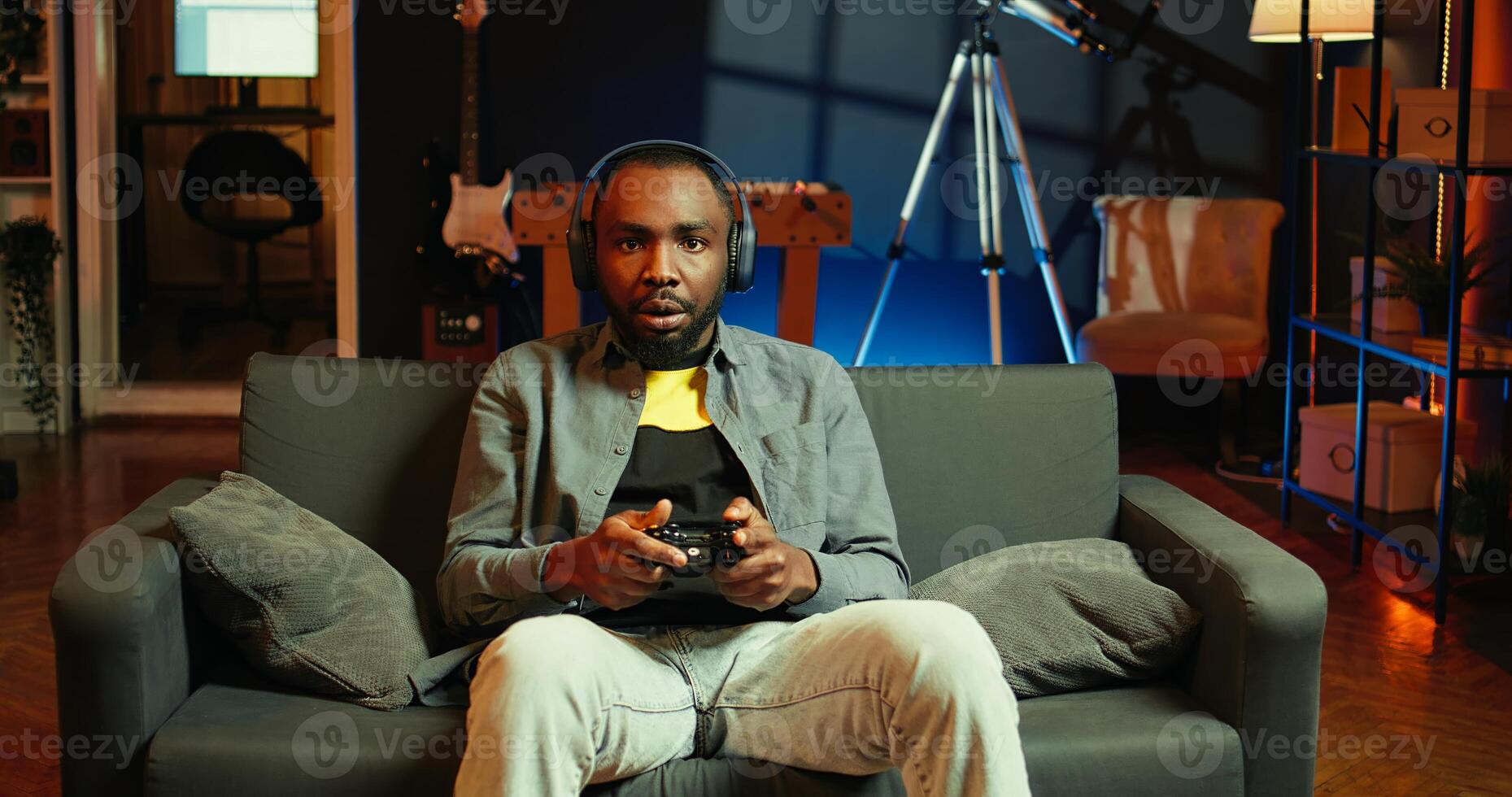 Cheerful man wearing headphones, celebrating after victory in videogame, neon lit apartment. Person on couch throws fists in air from excitement after winning game on console with controller, camera B photo