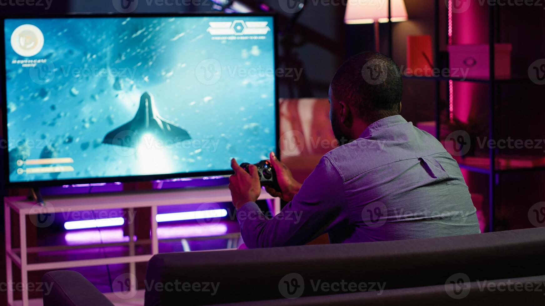 Gamer using controller and headphones to play first person shooter videogame on large smart TV. Man in home theatre using high tech gaming console gamepad to defeat foes, camera A photo