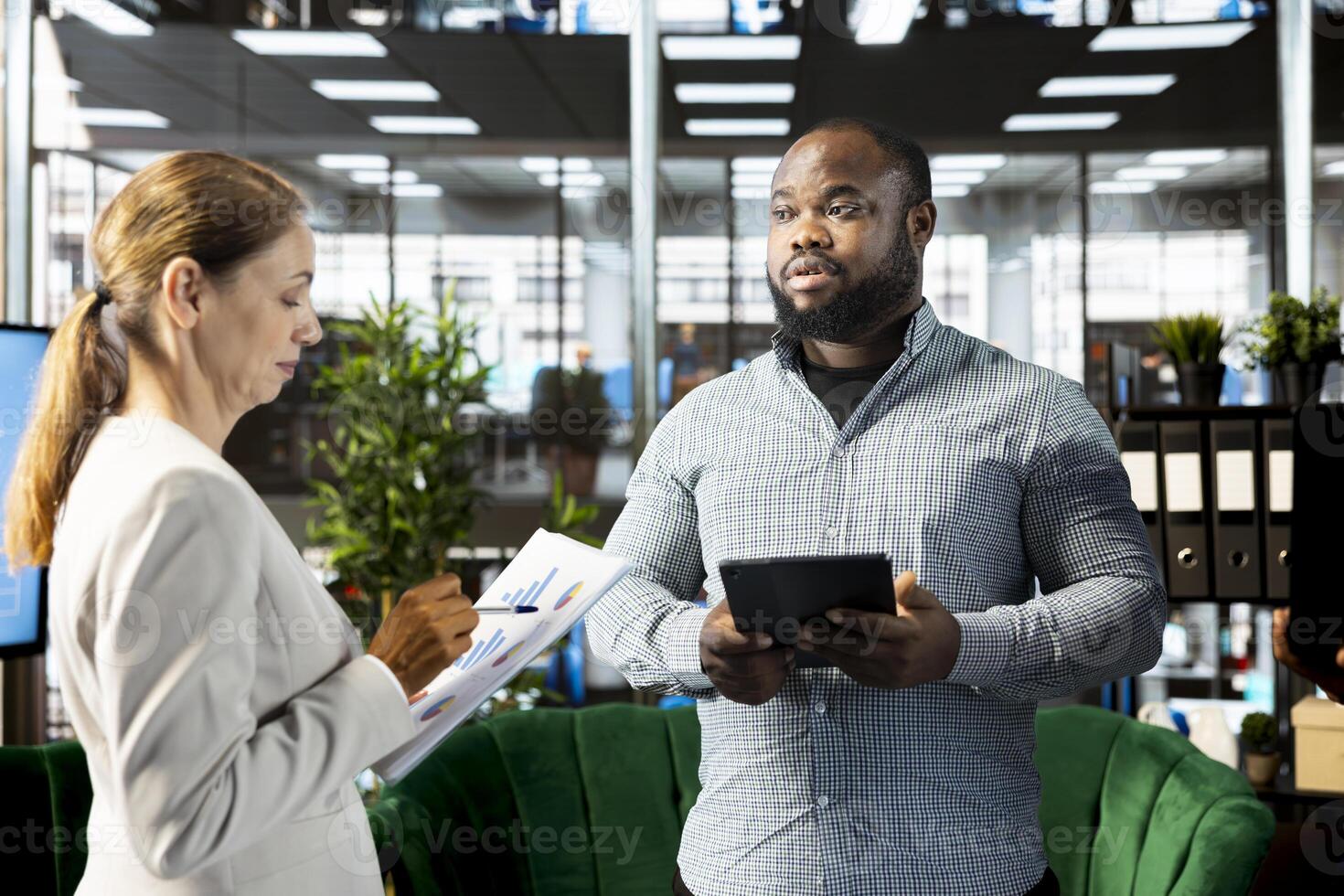 Employee presenting documents to manager holding tablet, showing him researching key data needed for finding business insights. Company executives analyzing data on device and paperwork photo