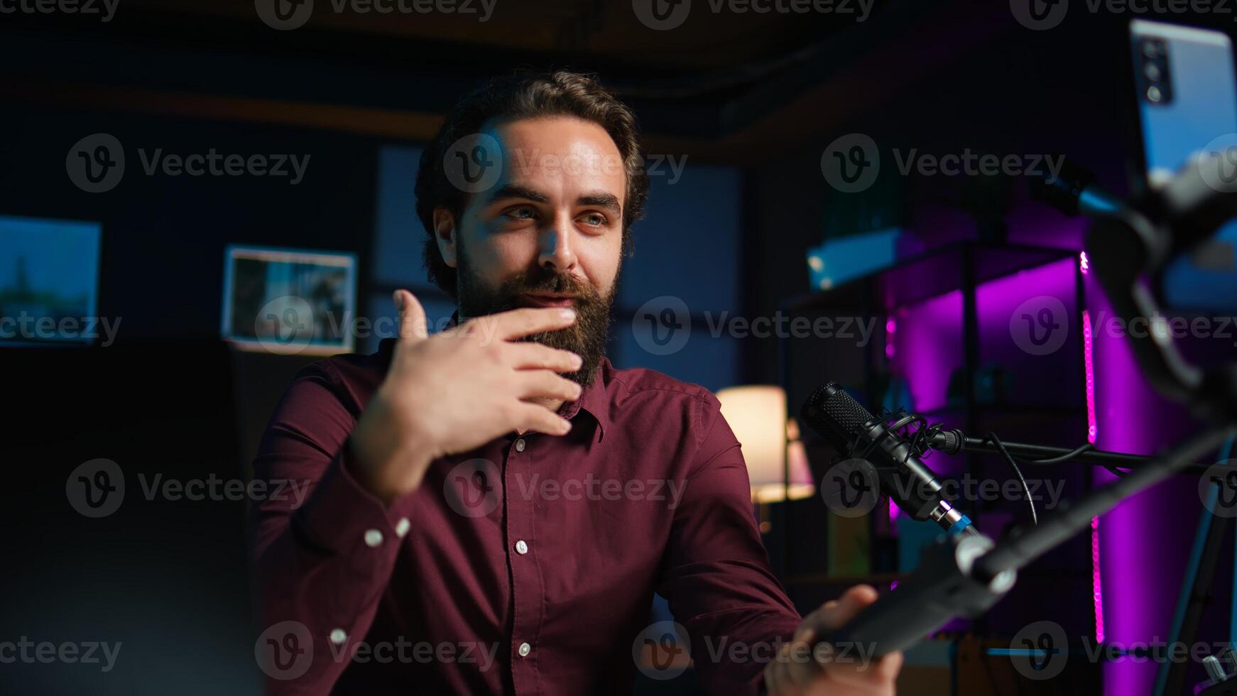 Happy influencer in home studio greeting audience, recording live broadcast using smartphone on selfie stick, close up. Man filming q and a session with mobile phone, answering questions, camera B photo