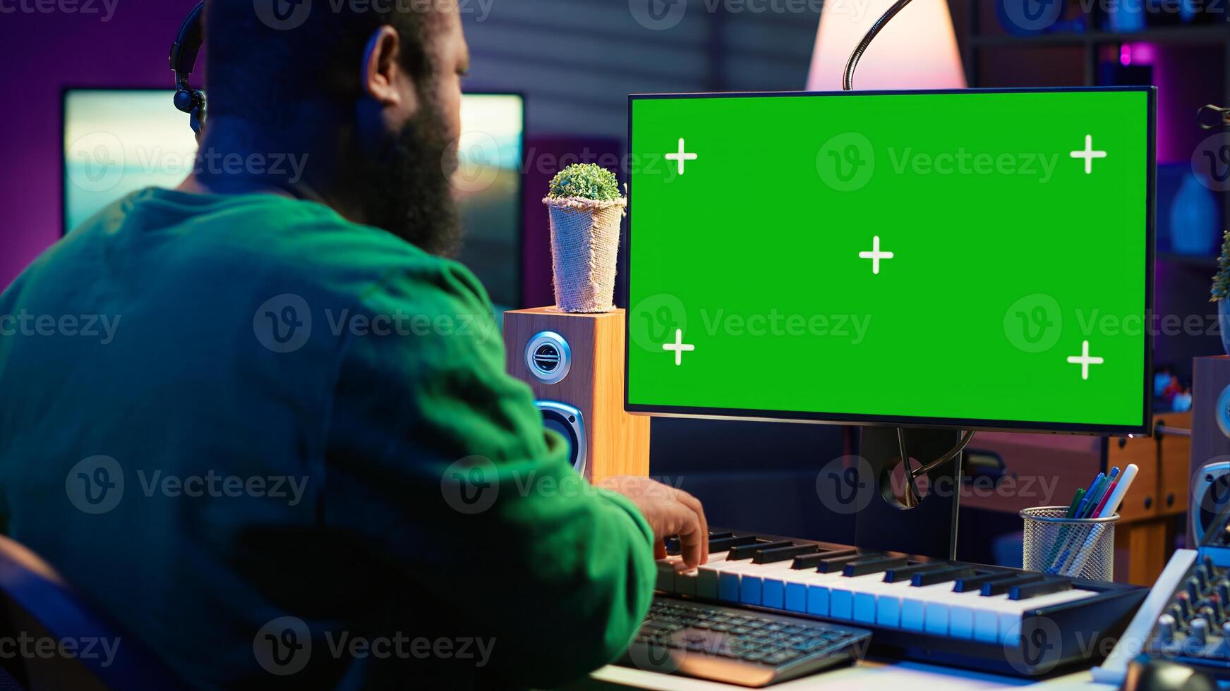 Musician operates on midi controller piano keys next to mockup screen, producing new songs with musical instruments and audio gear in home studio. Artist works with green screen. Camera A. photo