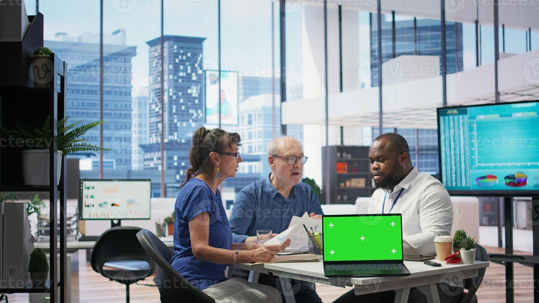 Banker guiding senior couple in choosing the right retirement plan, having a green screen on laptop in modern office. Elderly people consulting insurance agent about financial options. Camera B. photo