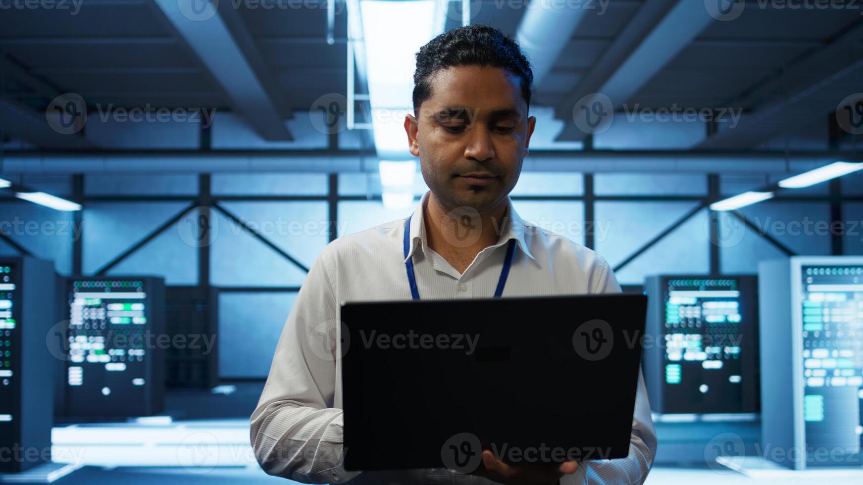 System administrator in data center using notebook to design, implement and manage gear, ensuring optimal performance and reliability. Indian man in server room coding on laptop, camera B photo