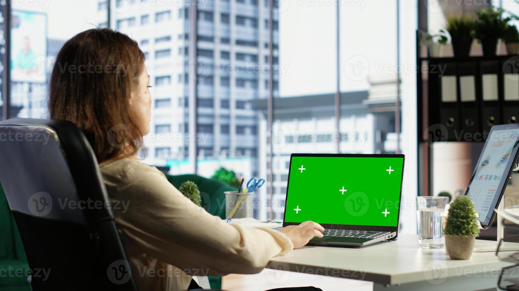 Young business entrepreneur looking at green screen mockup on her gadgets, working in modern urban corporate office. Female leader developing a strategy to increase funding for success. Camera B. photo
