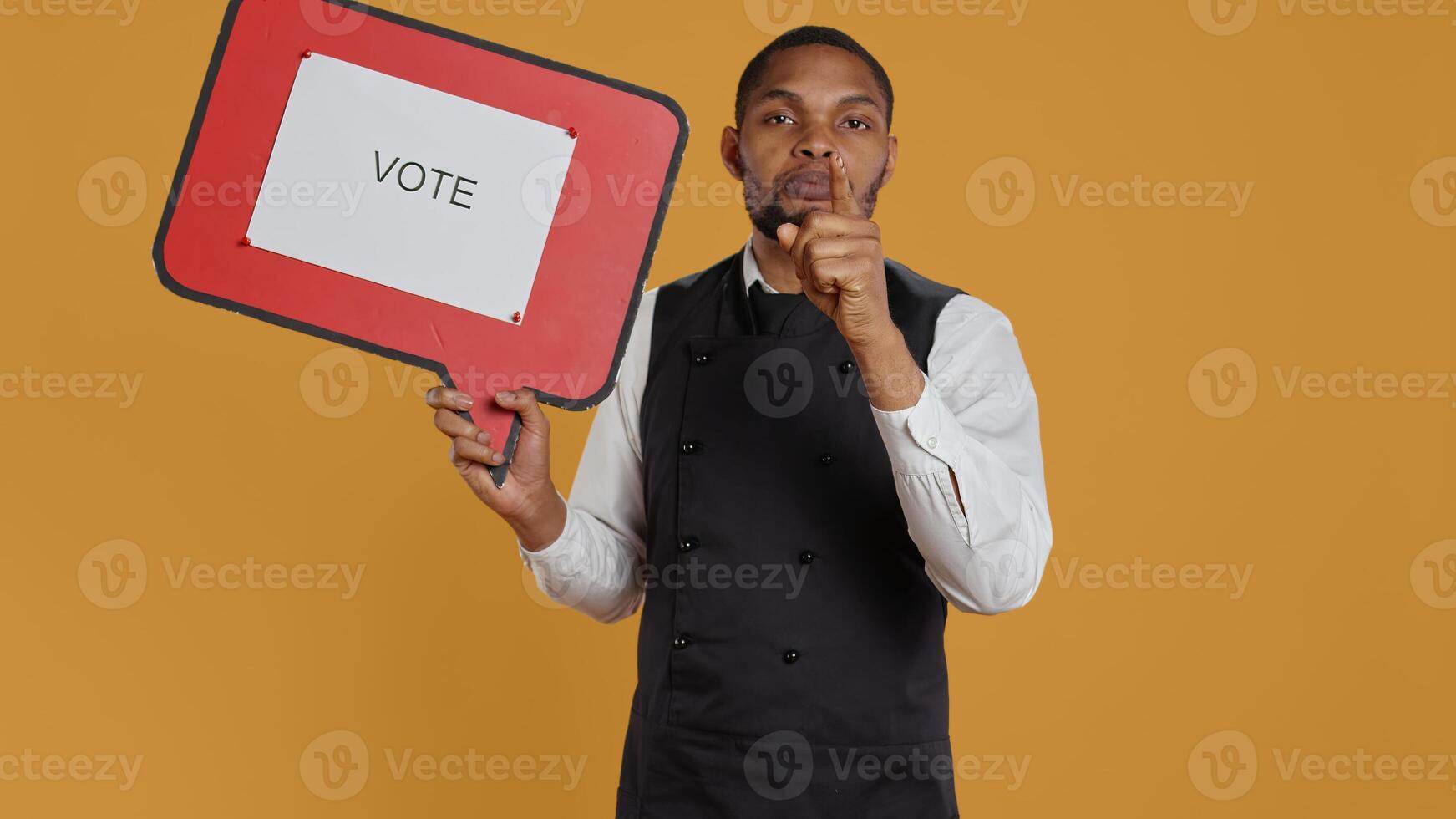 Catering employee showing a message to vote on red speech bubble, presenting the cardboard icon to do a commercial for politics. Waiter in uniform works on customer service. Camera B. photo