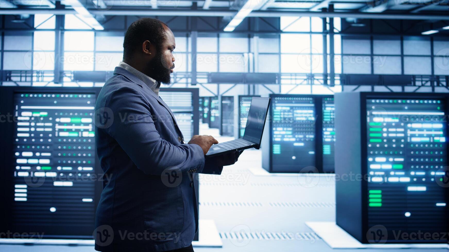 African american man walking in high tech facility, using laptop to patch network issues leading to slow data transfer rates. Data center worker fixing errors affecting supercomputers, camera B photo