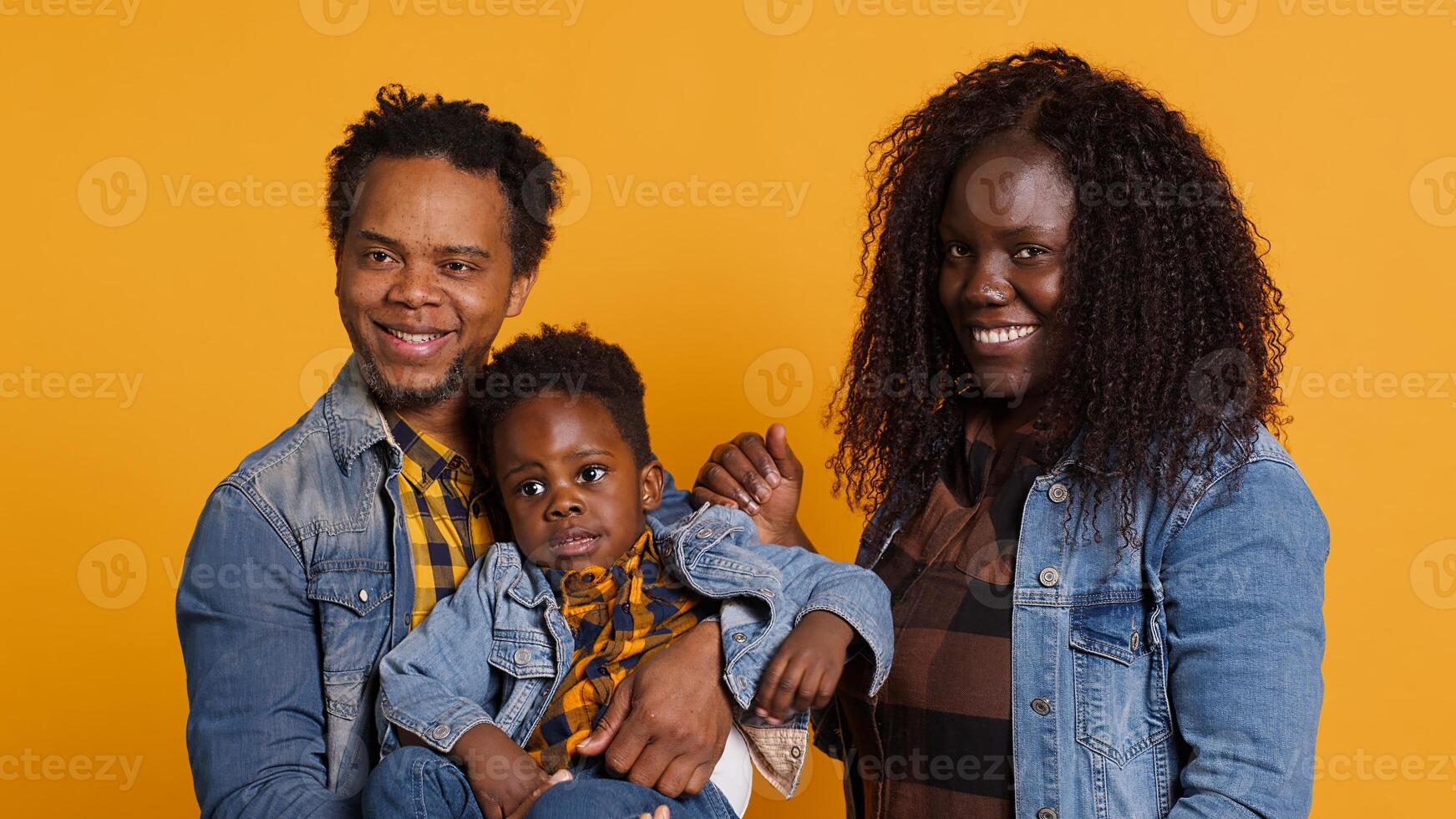African american family smiling against yellow background in studio, posing with love and sincere gratitude. Positive optimistic mom and dad holding their baby boy, enjoying time together. Camera A. photo