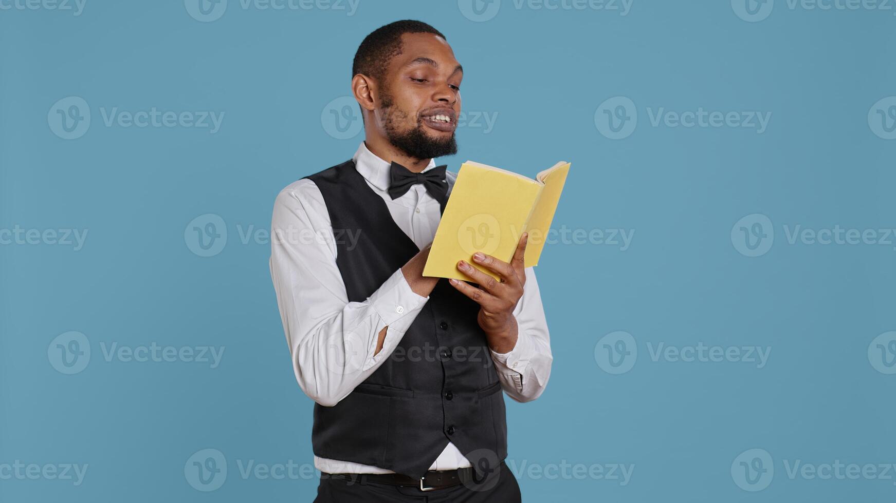 Young doorman hotelier reading a book on his work break in studio, hotel concierge in tuxedo against blue background. Bellhop resort personnel enjoys literature and lecture. Camera B. photo