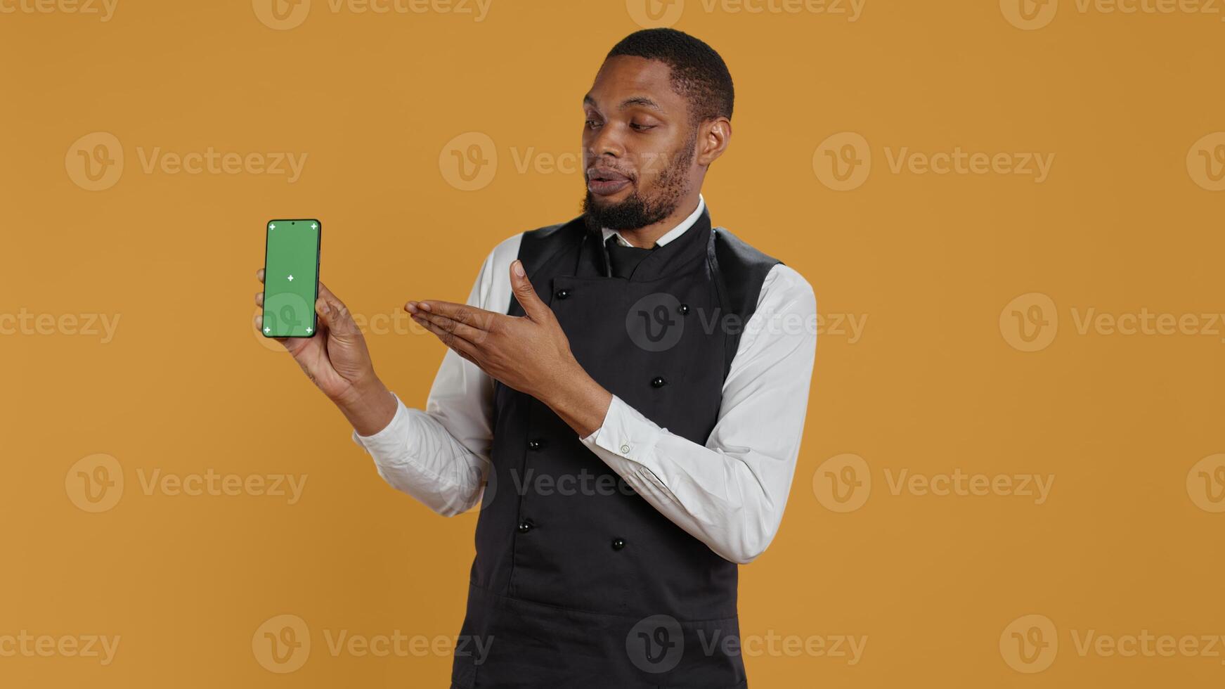 Restaurant staff showing a smartphone with green screen in studio, prepared to serve food orders in service oriented environment. Waiter with apron and uniform serving clients at tables. Camera B. photo