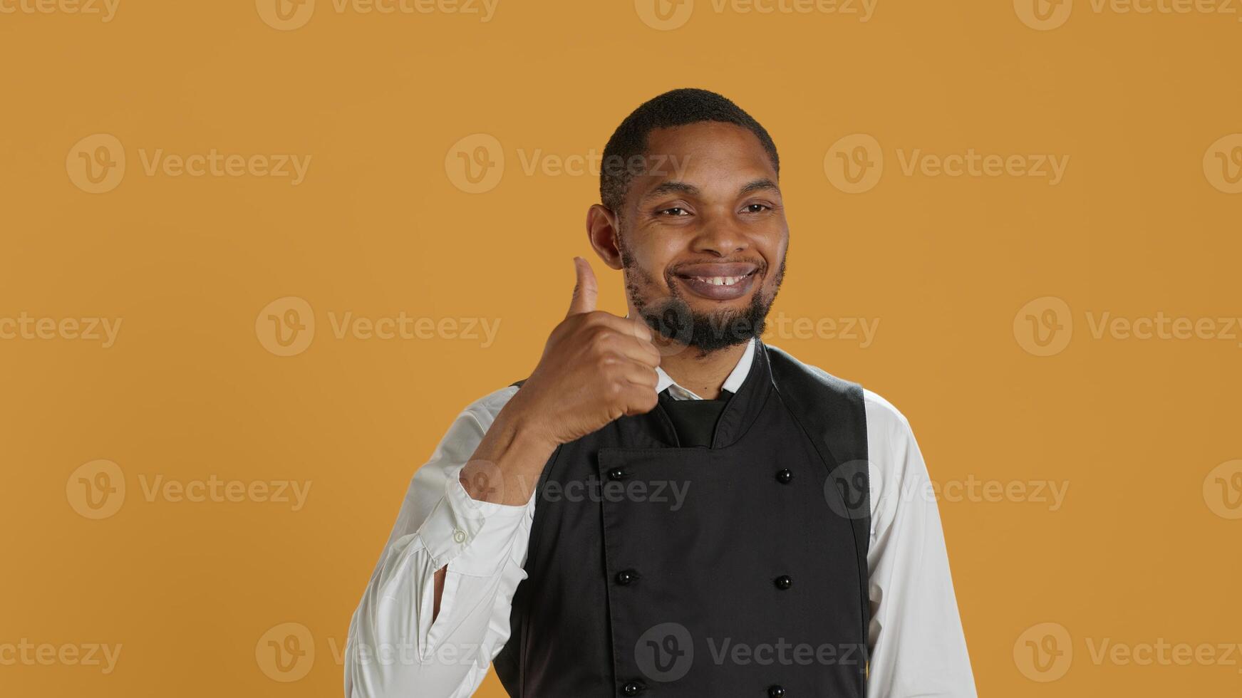 Waiter smiling and showing a thumbs up symbol against yellow background, presenting his satisfaction and approval with a positive like sign. Cheerful staff gives okay gesture. Camera A. photo