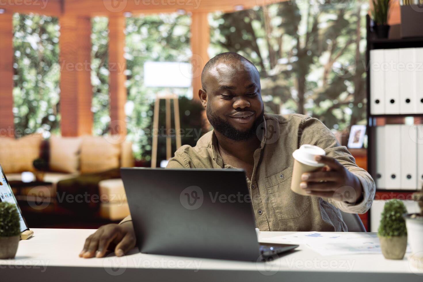Business owner working remotely on a laptop and drinking coffee, enjoying his home based setup for enterprise activity. Handling operations online, showcasing successful telecommuting. photo