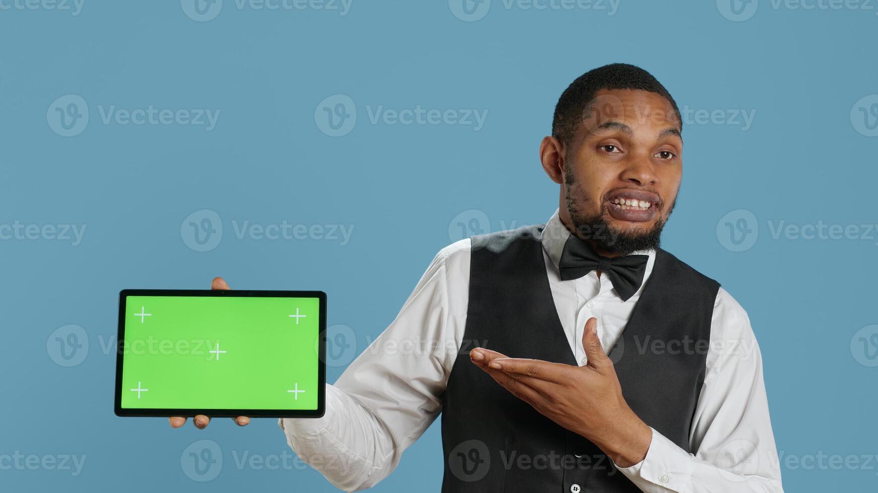 Doorkeeper bellboy poses with green screen on tablet at the hotel reception, working in hospitality industry and greeting customers. Lodging resort personnel shows chroma key. Camera A. photo