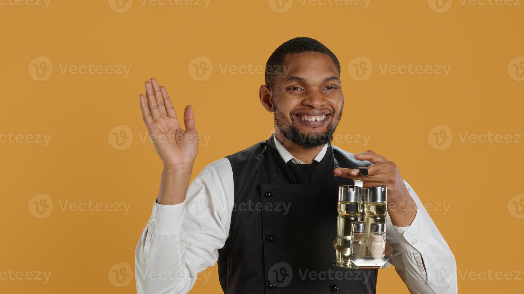 Happy friendly waiter waving hello and welcoming clients at table, holding a cruet set with condiments ready to serve customers. Cheerful lovely staff greeting people at restaurant. Camera A. photo