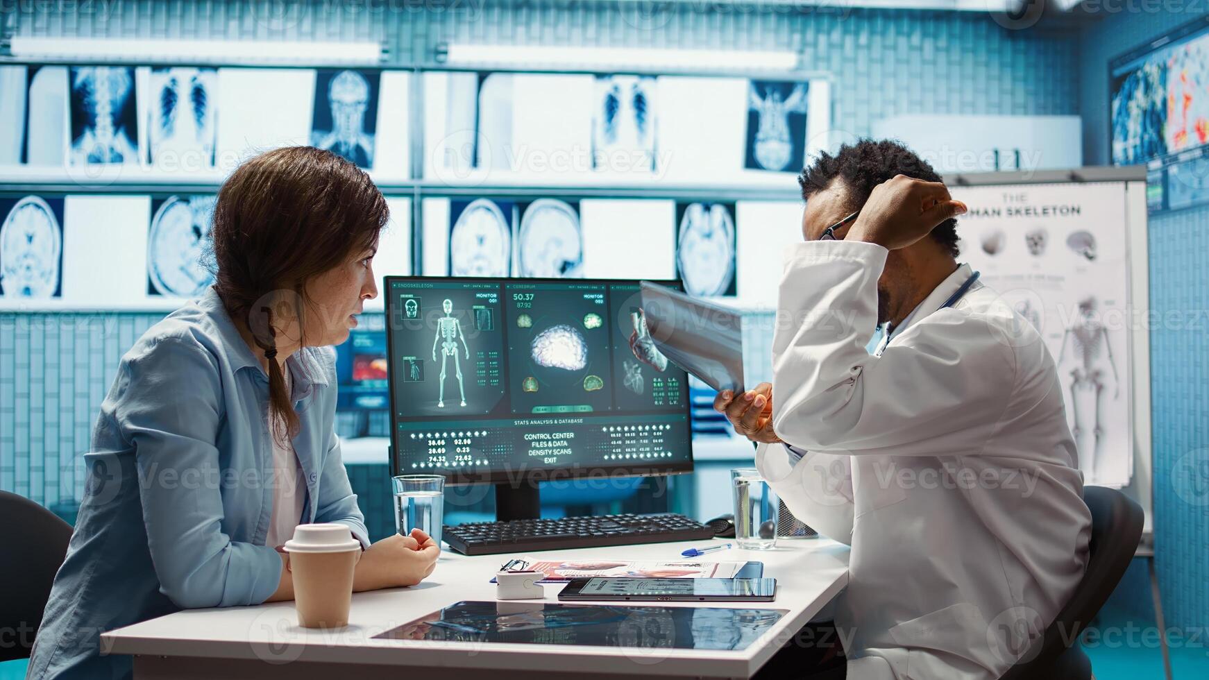 African american physician presenting the x ray diagnostics results to a patient, examining the radiography scans in a medical cabinet. Doctor recommending medicine for wellness. Camera B. photo