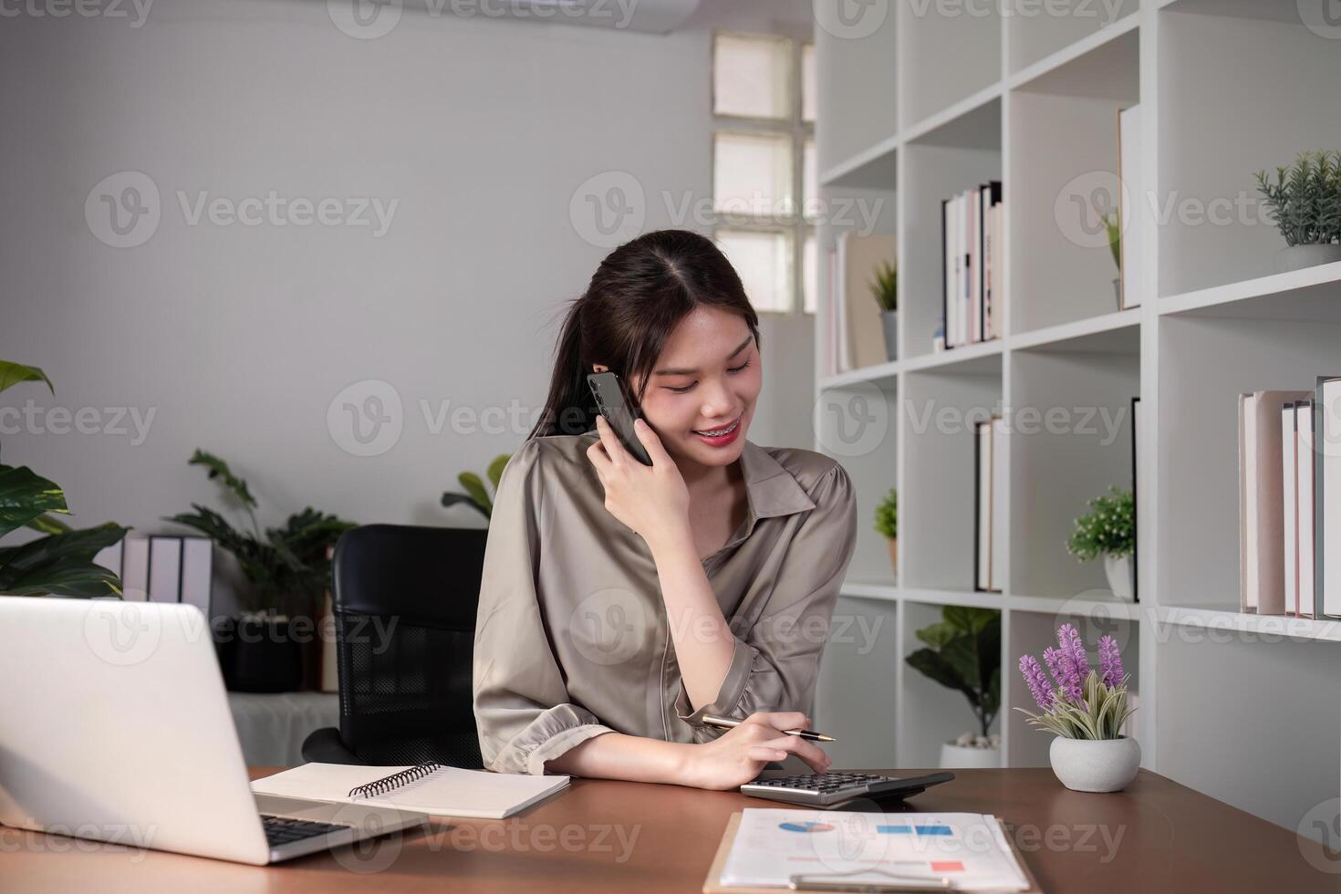 Young Asian business woman sits on the phone in an online business meeting using a laptop in a modern home office decorated with shady green plants. photo
