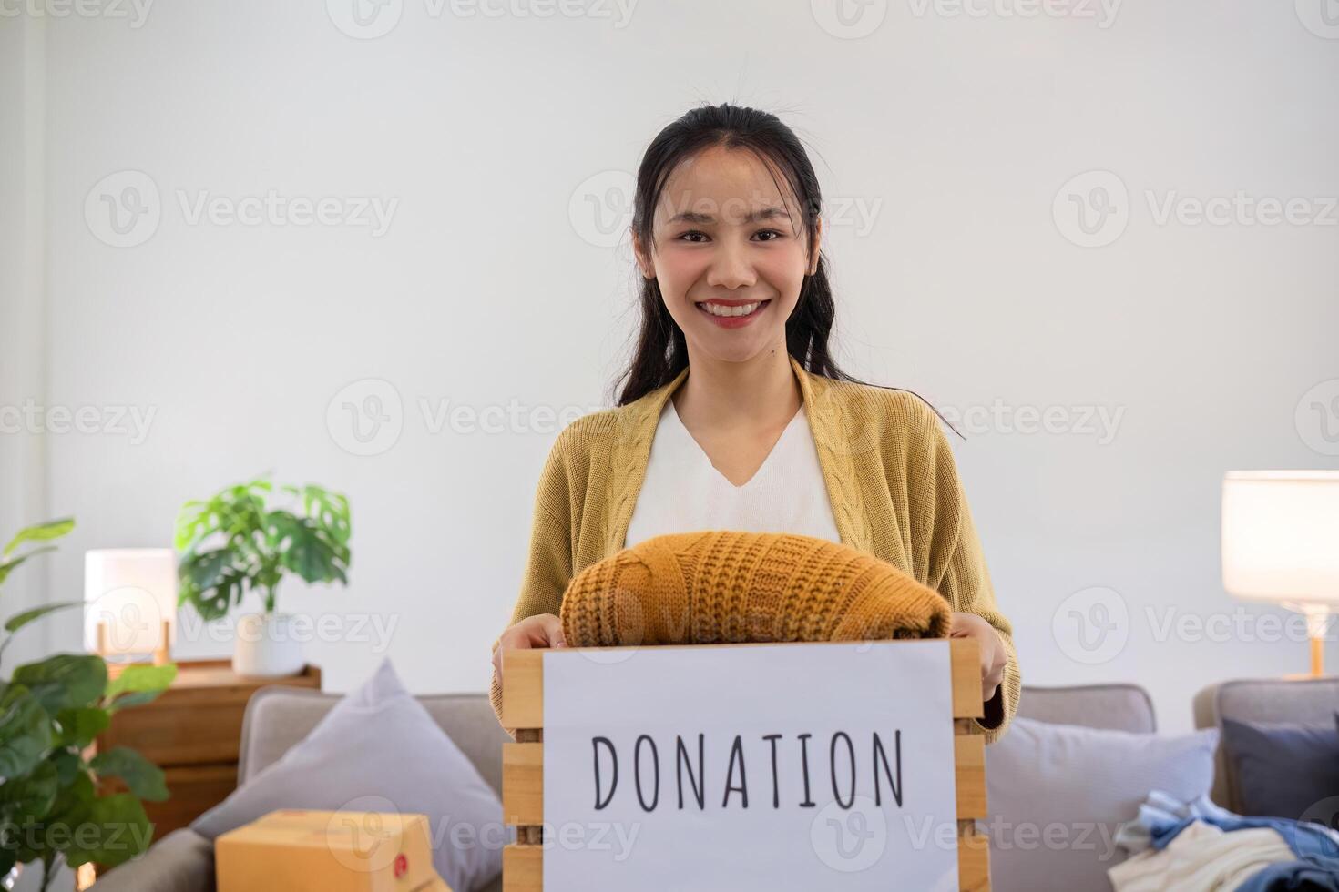 A cute Asian woman stands holding a box, sorting unwanted clothes for donation. photo