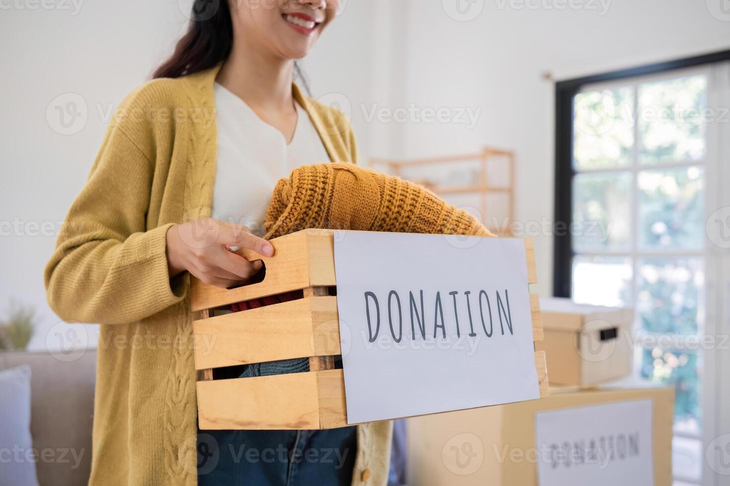 A cute Asian woman stands holding a box, sorting unwanted clothes for donation. photo