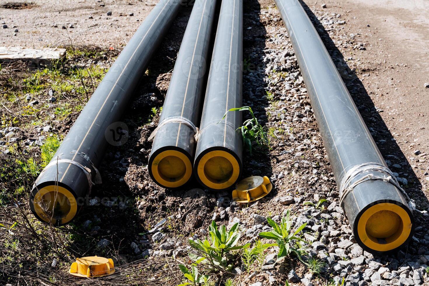 Closeup of large industrial metal pipes with yellow ends resting on gravel and surrounded by vegetation on a construction site. Sunlight illuminating the engineering infrastructure project photo
