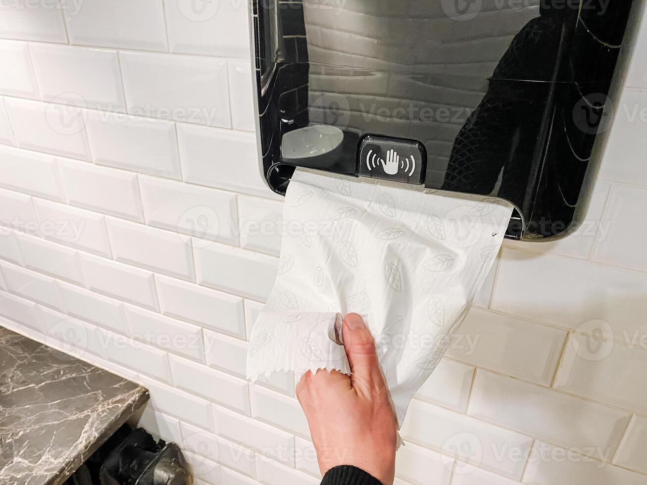 A hand gestures to the paper towel dispenser in front of a cabinetry window. photo