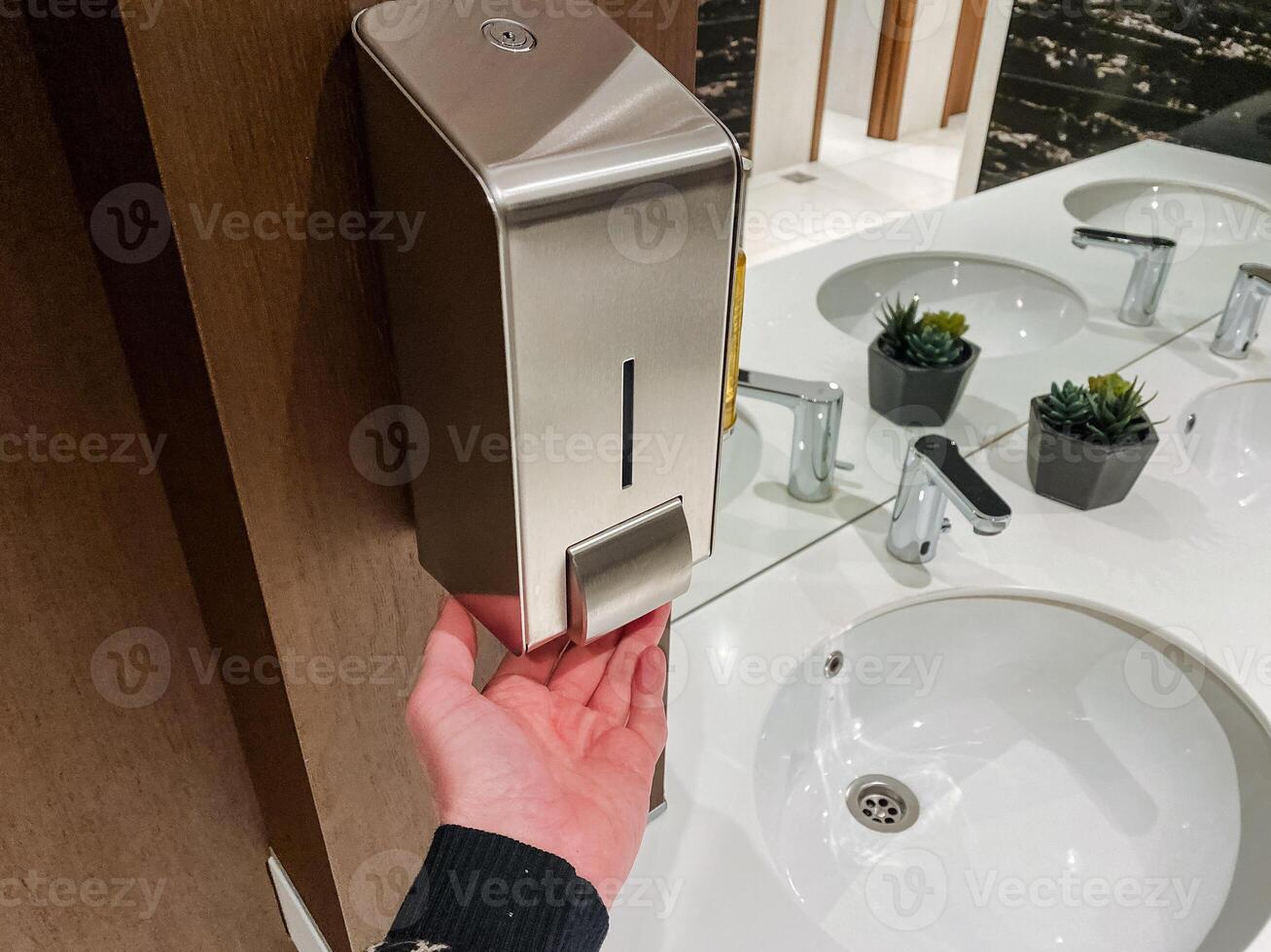 A person using a soap dispenser in a bathroom with hardwood flooring and wood stain on the cabinetry of a building photo