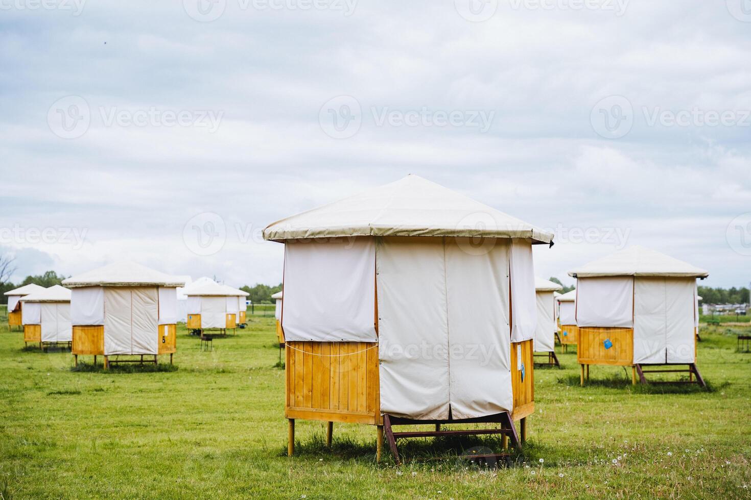 In a rural setting, community structures stand in an open grass field. There are temporary wooden and canvas shelters under a cloudy sky, creating a serene and picturesque scene photo
