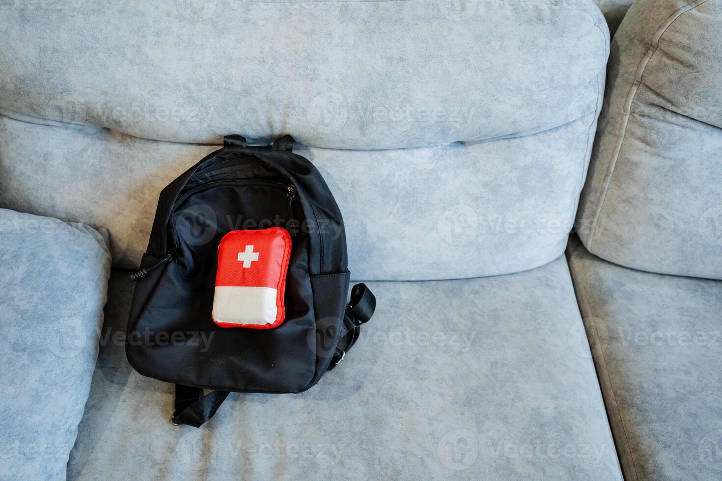A first aid kit is on the backpack, a black backpack on the couch, a travel bag, a small red box with medicines. photo