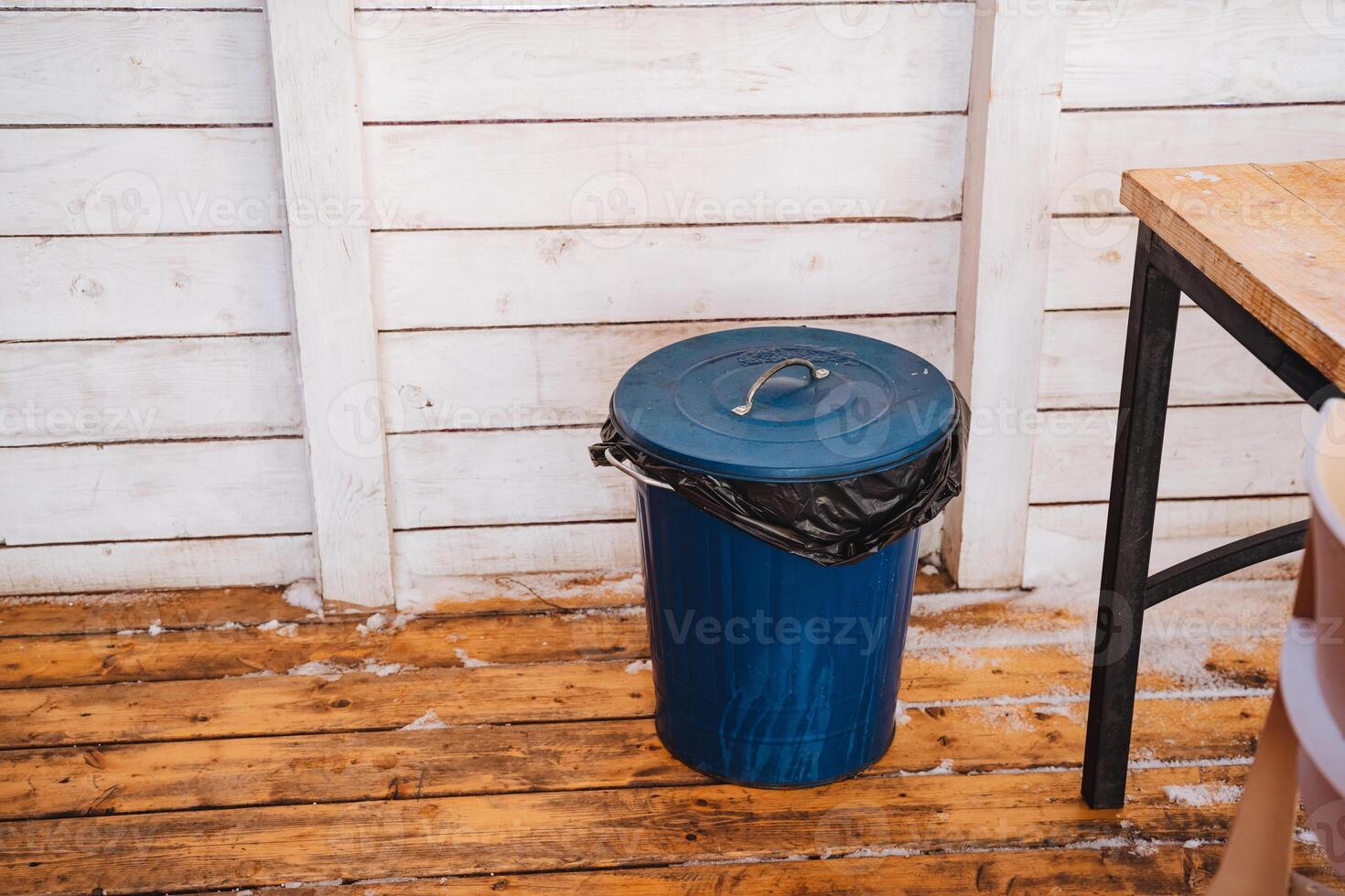 A vibrant blue trash can can be seen sitting on a beautifully polished wooden floor, placed next to a rustic style table that enhances the decor and overall ambiance of the entire space photo