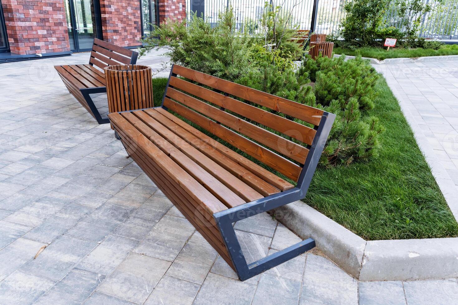 In a park, there is a line of wooden benches with a brick building in the background. The setting includes green grass and a wooden structure that complements the outdoor scenery photo