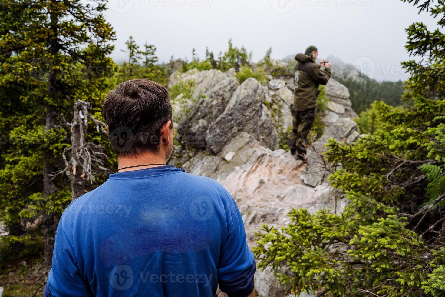 A man dressed in a vibrant blue shirt stands with confidence atop a rocky hill, taking a serene moment to appreciate the breathtaking natural landscape that surrounds him completely photo