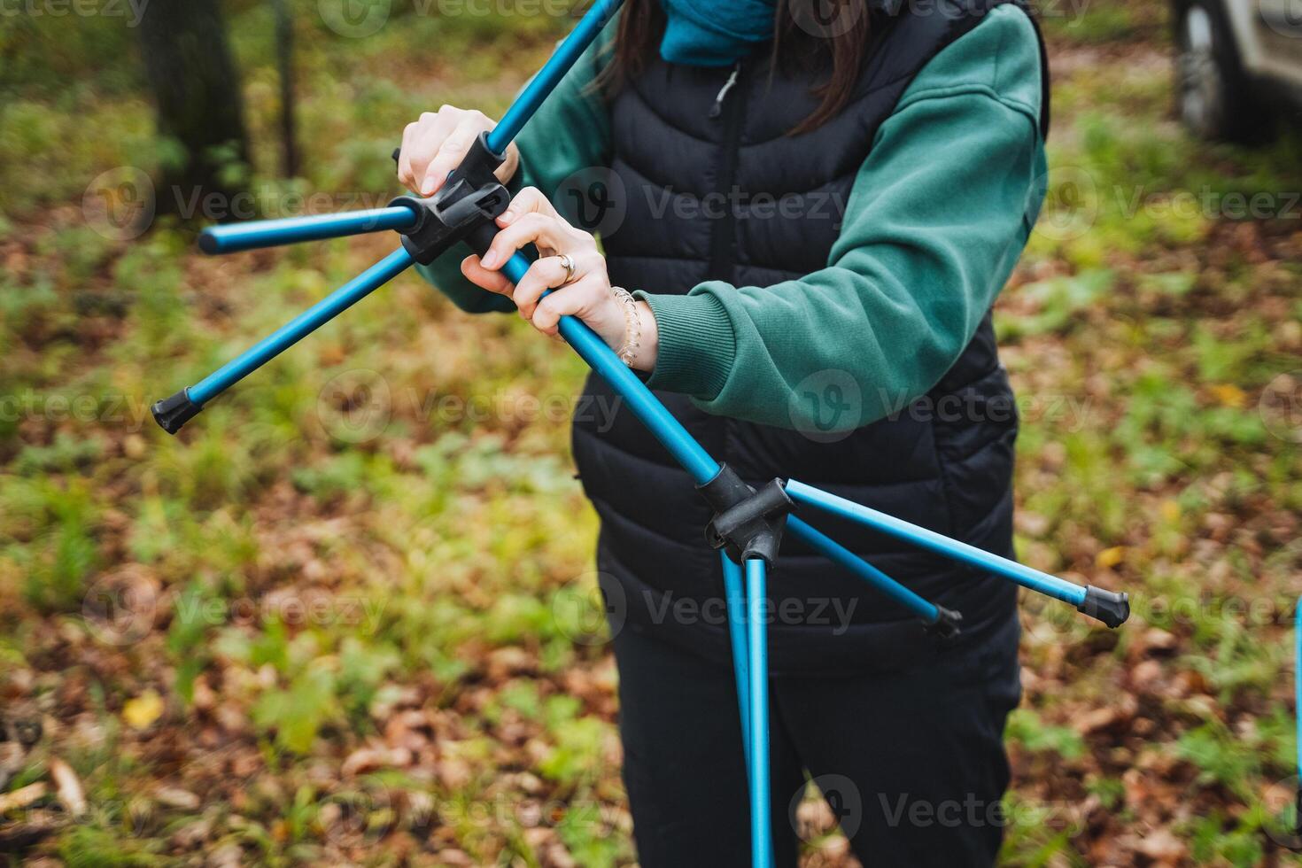 A confident woman stands with pride in a serene wooded area, firmly holding a blue tripod, fully prepared for an adventure amidst the lush greenery and towering majestic trees photo