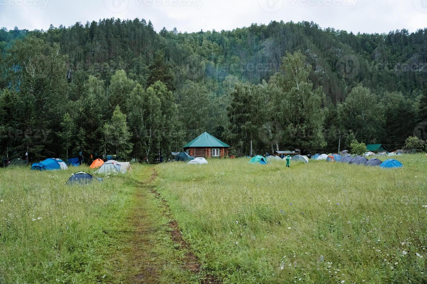 The expansive field is sprinkled with tents and a distant house, set under a clear sky amidst lush greenery and trees, creating a picturesque natural landscape of grassland photo