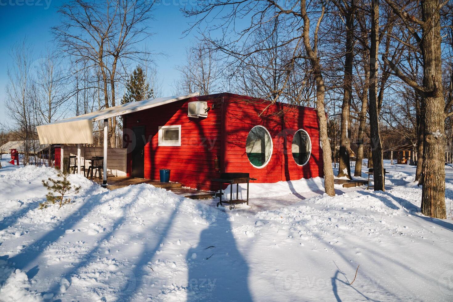 In the heart of a pristine, snowy forest, surrounded by tall trees and delicate twigs, stands a charming red house that offers a warm shelter amidst the cold winter landscape photo