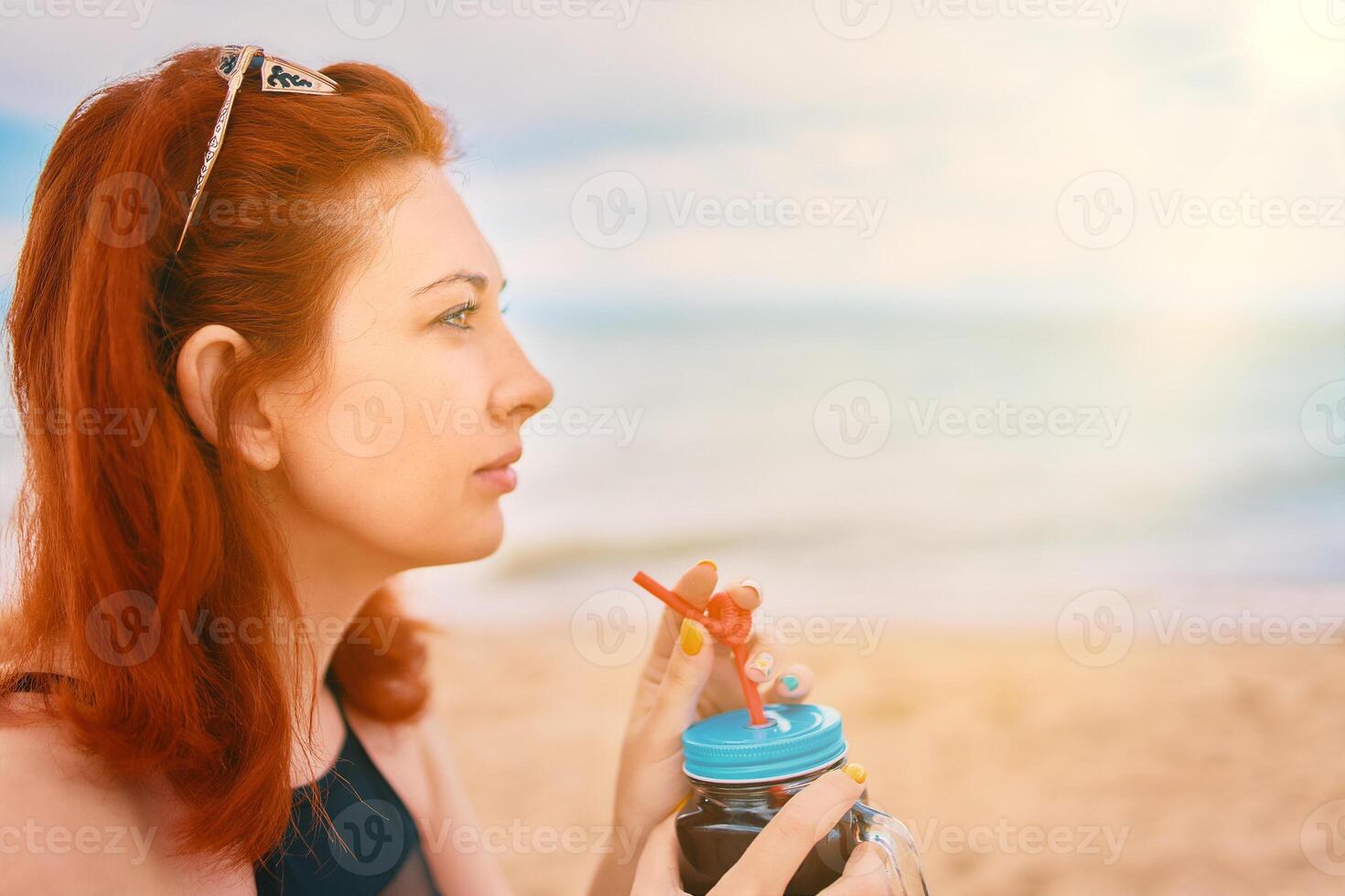 Red-haired girl in a swimsuit is sitting on the beach with a cocktail and look away. Calm woman on vacation. Holidays at the sea. Vintage sunglasses on the head of a having a rest lady. photo