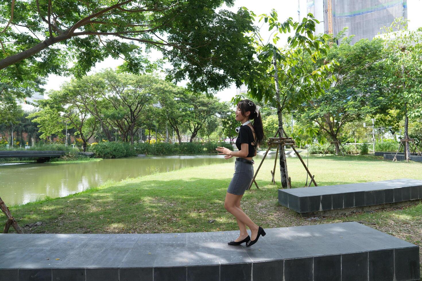 A young woman enjoying a moment of freedom, posing playfully on a green lawn in a sunny urban park, surrounded by trees and pathways, exuding a sense photo