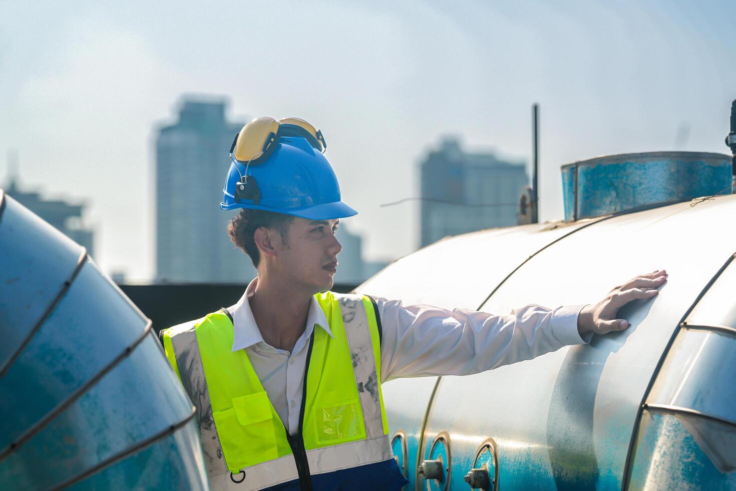 Engineer with safety gear inspects large industrial water tanks on a city rooftop against a skyline. photo