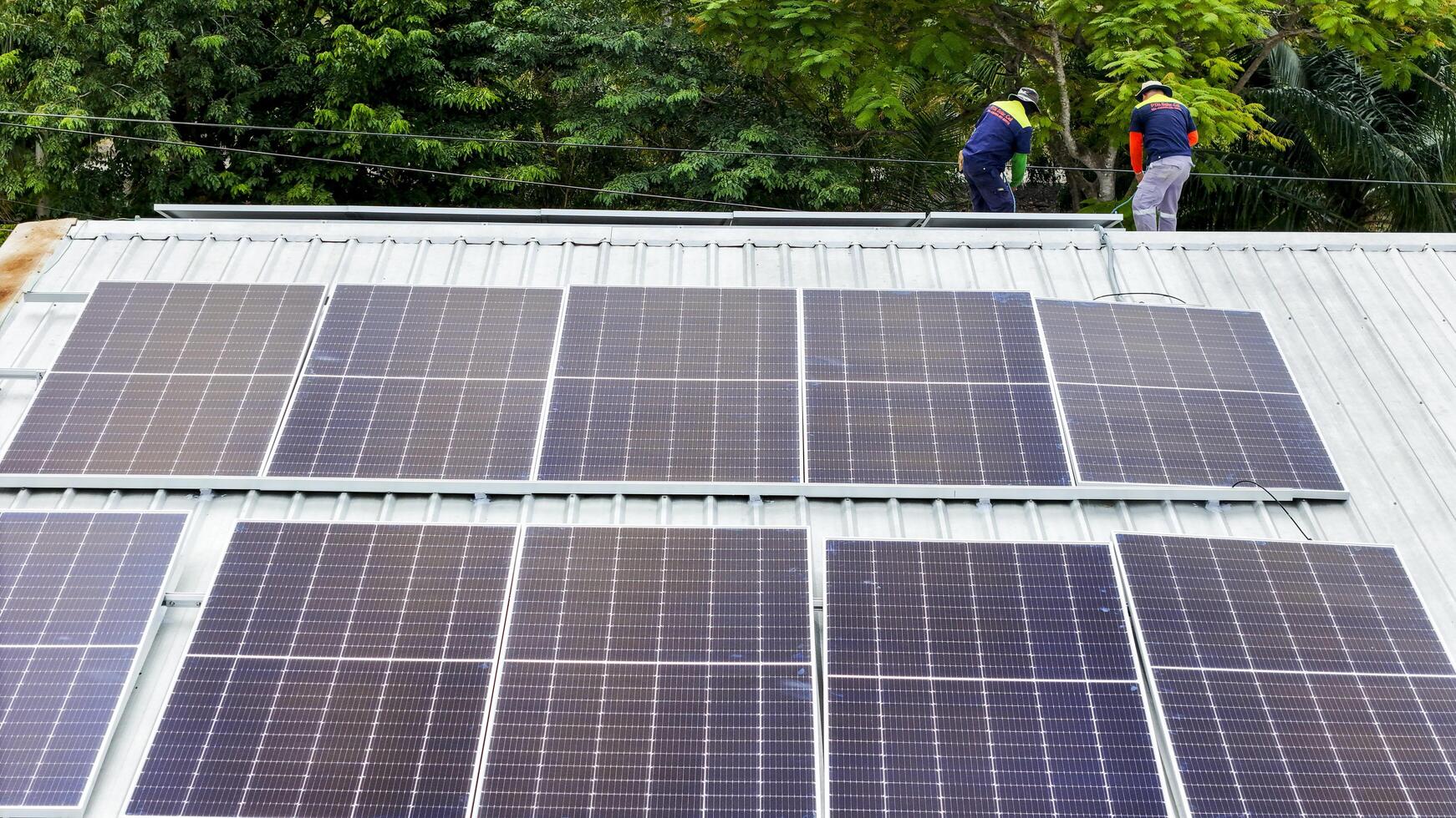 Solar panels installed on a corrugated metal roof, with technicians working amidst green trees, symbolizing the harmony between renewable energy and nature in a modern setting. photo