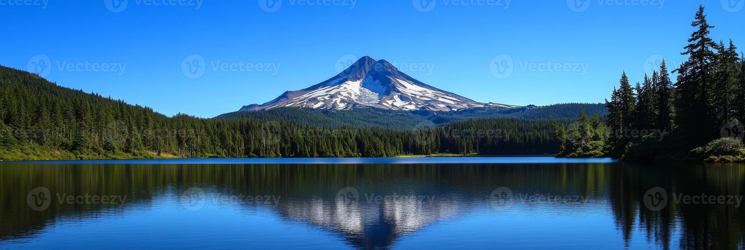 A peacefully tranquil mountain lake mirrors the stunning, rocky peaks and vibrant greenery surrounding it, with a clear blue sky overhead enhancing the already picturesque landscape that captivates photo