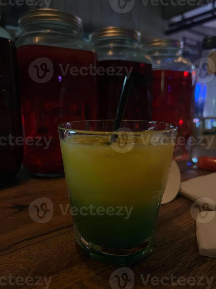 A vibrant cocktail sits on a rustic wooden table, featuring a colorful gradient from yellow to green. Various jars in the background set a lively atmosphere for the drink. photo