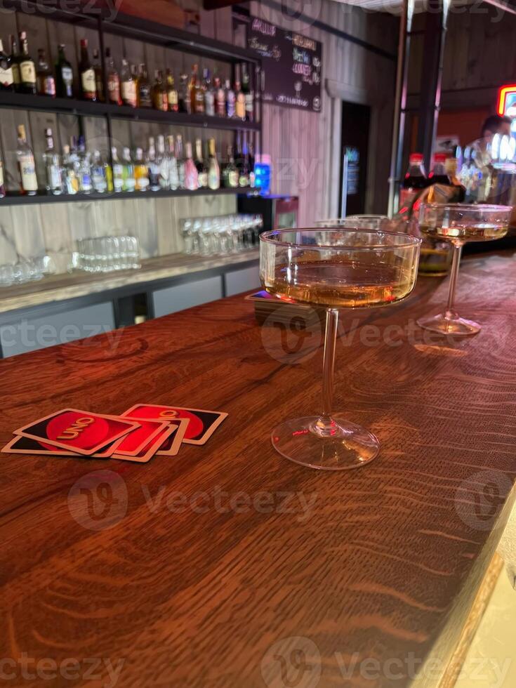 Two elegant cocktail glasses rest on a polished wooden bar, with UNO cards nearby. The vibrant backdrop suggests an inviting social setting perfect for games photo