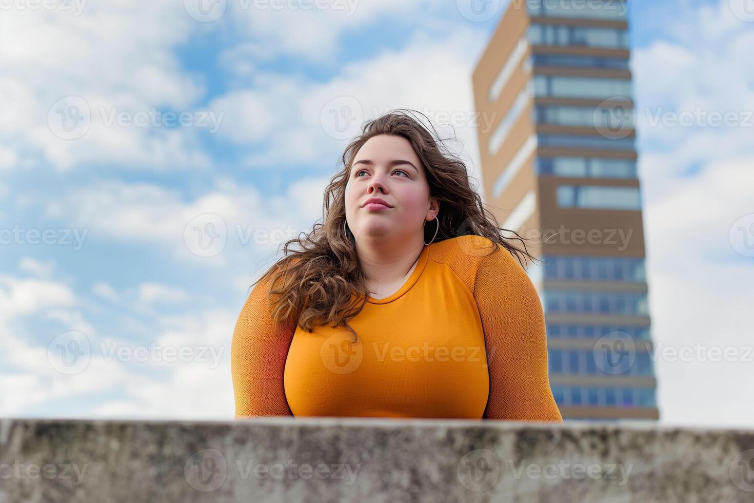 A plus size young woman stands confidently outdoors, looking toward the sky with modern buildings behind her photo