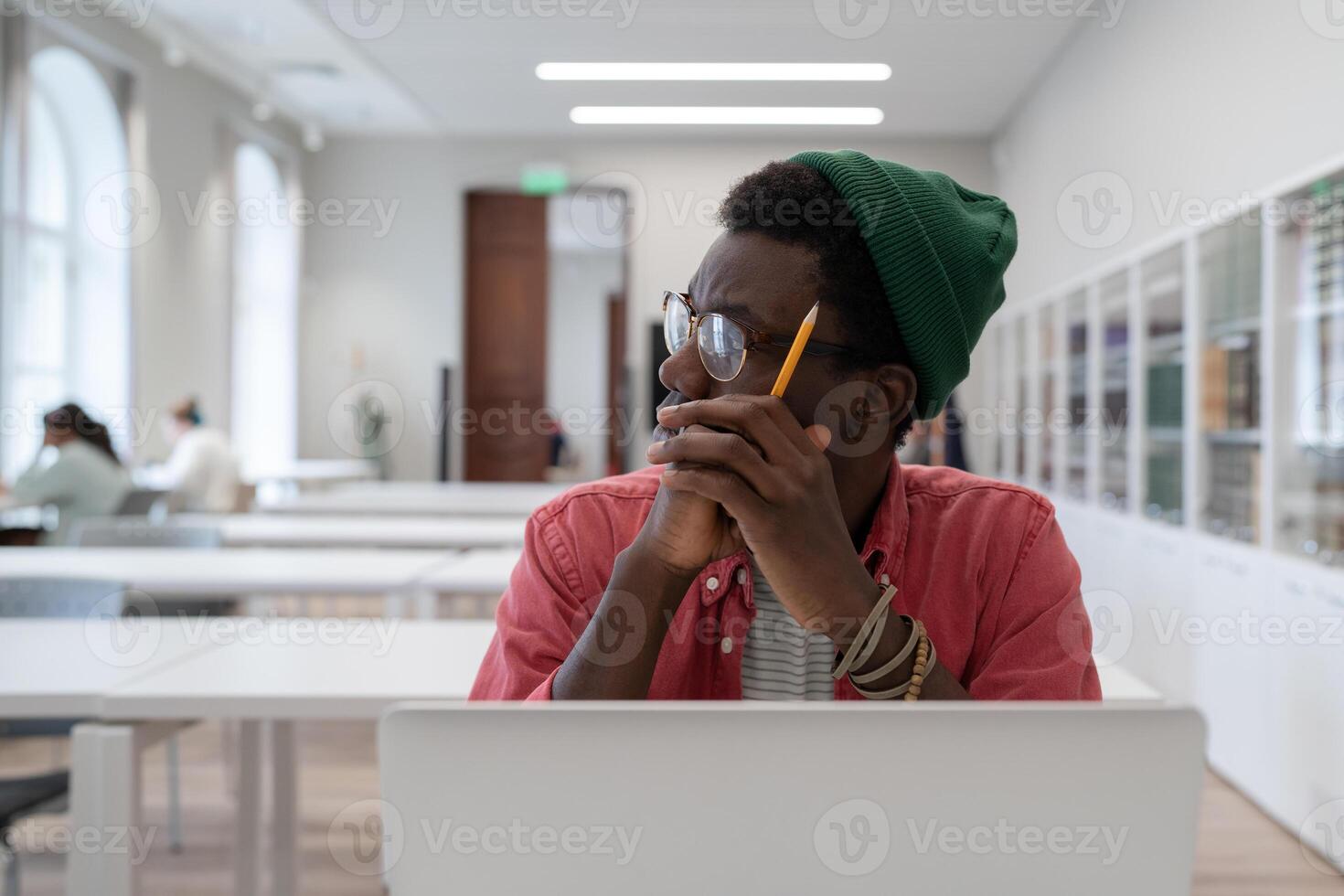 African student guy distracted from study, sitting at table with laptop in library and looking aside photo