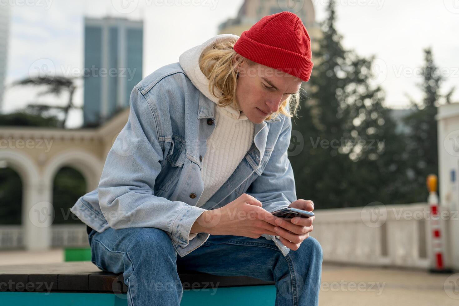 Pensive focused guy in red hat sitting outdoors, searching info in mobile phone, messaging chatting. photo