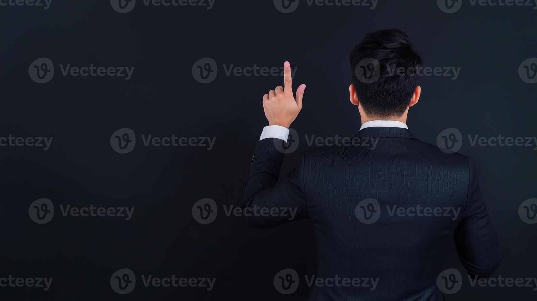 Man in a suit pointing upward with one finger, standing against a dark background. Rear view showing professional attire and gesture indicating direction or emphasis. photo