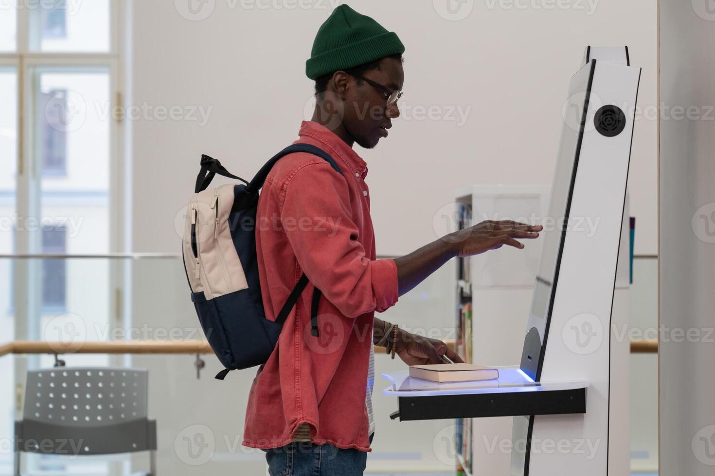 Young African student guy standing in library using self-service terminal to borrow or return books photo
