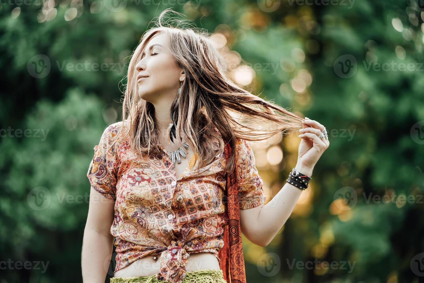 Beautiful young woman wearing bohostyle clothes posing in the rays of the evening sun, sunset. Boho style fashion, female wearing silver jewelry having fun in park outdoors. photo