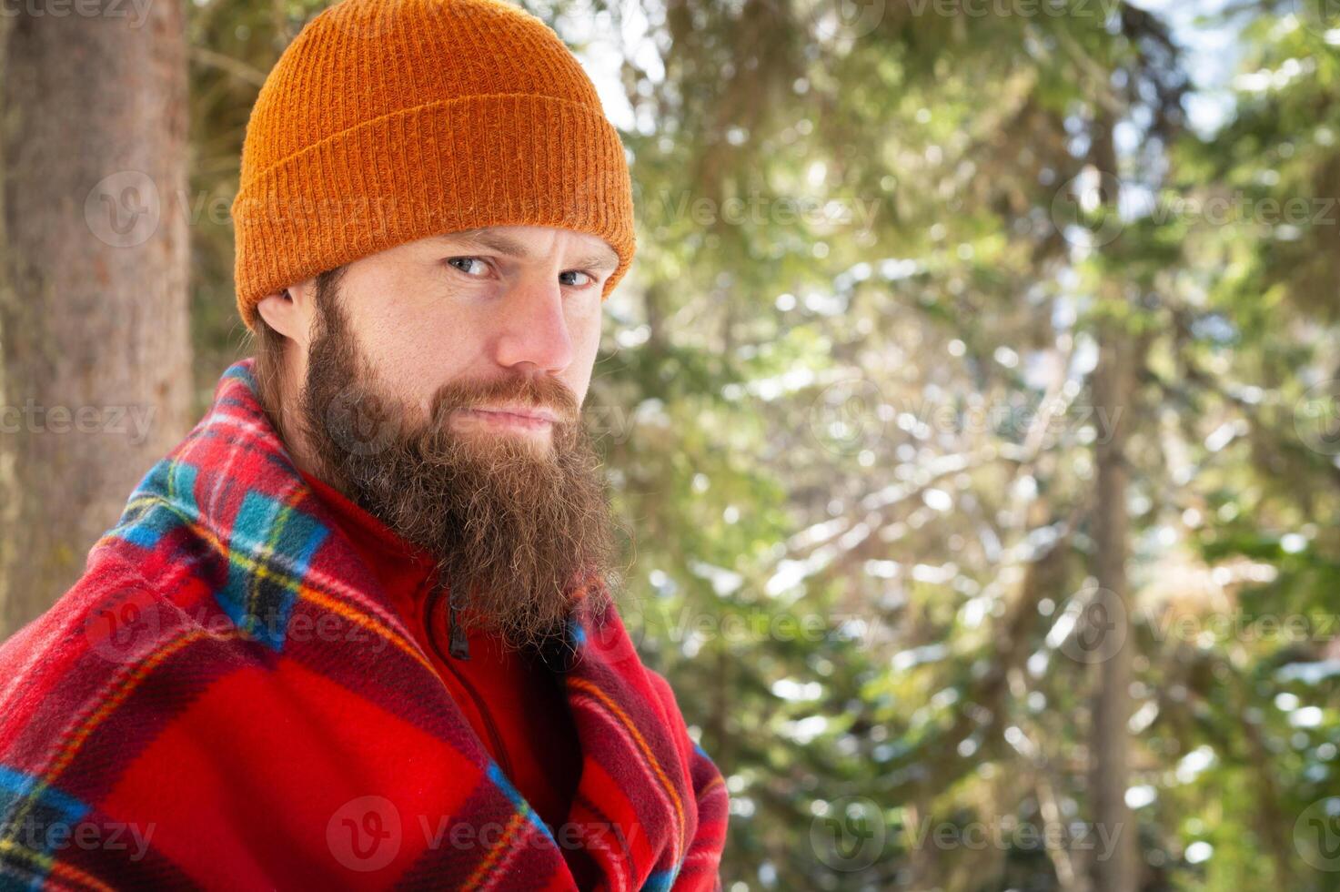 Bearded man smiling while standing among lush green forest with happy expression in checkered red blanket photo