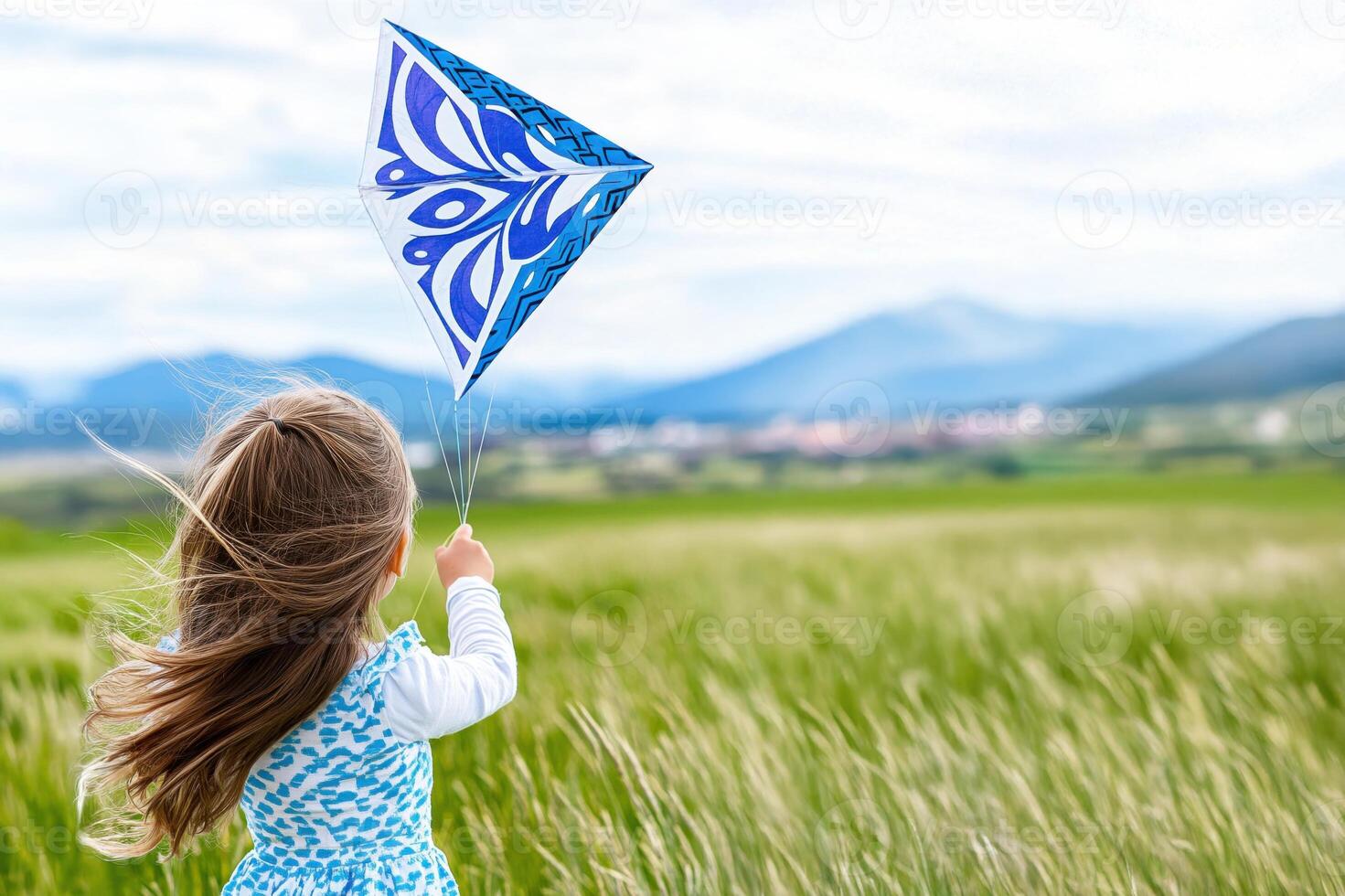 Young girl joyfully flying a blue kite in a green field against a backdrop of mountains on a sunny day photo